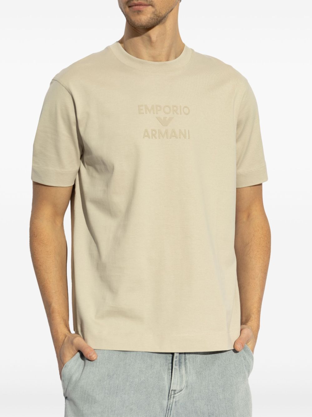 Man wearing Emporio Armani beige crew neck T-shirt with embossed logo, short sleeves, straight hem, paired with jeans.