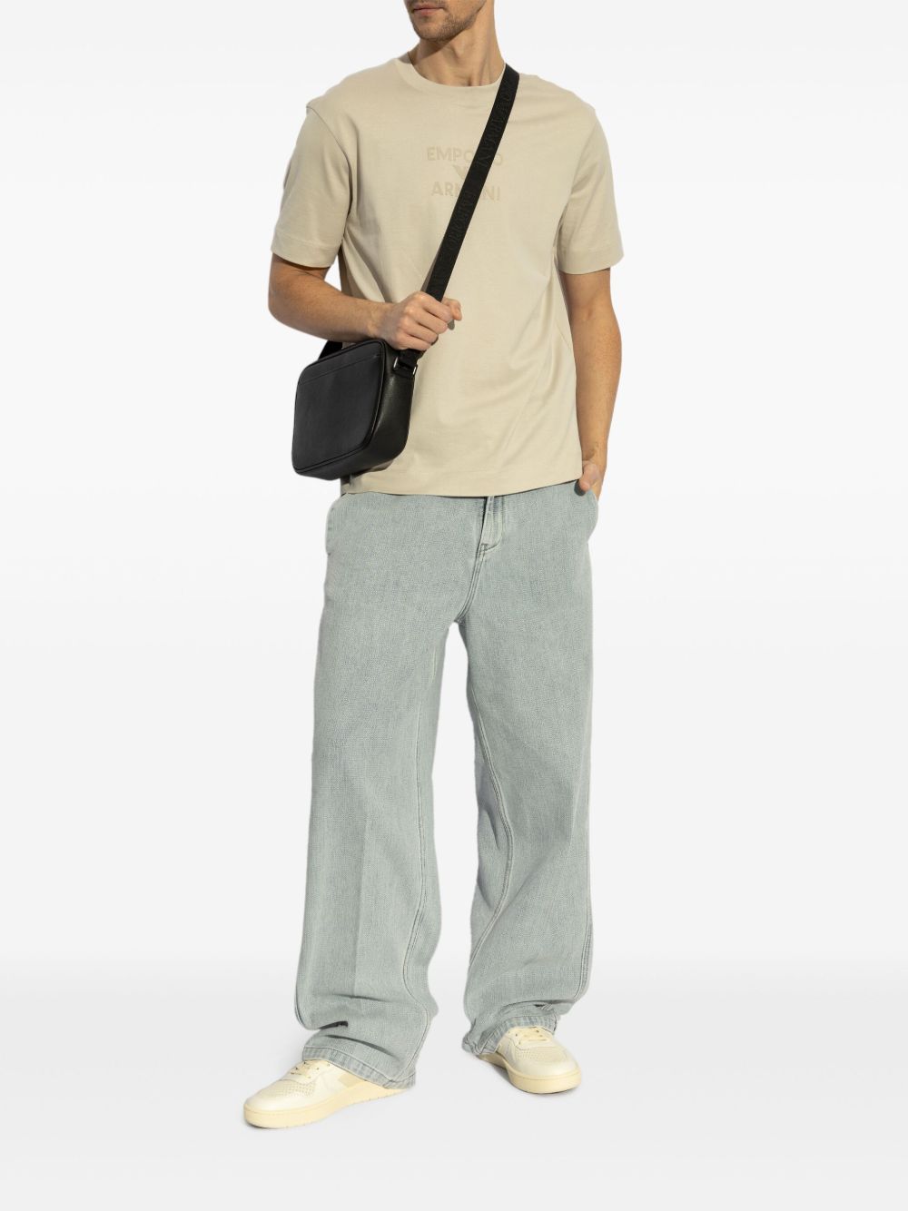 Model wearing beige Emporio Armani T-shirt with embossed logo, paired with light blue jeans and black shoulder bag.