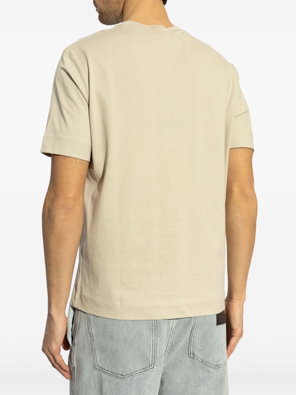 Back view of a beige Emporio Armani logo printed T-shirt with short sleeves, worn by a model in light denim jeans.